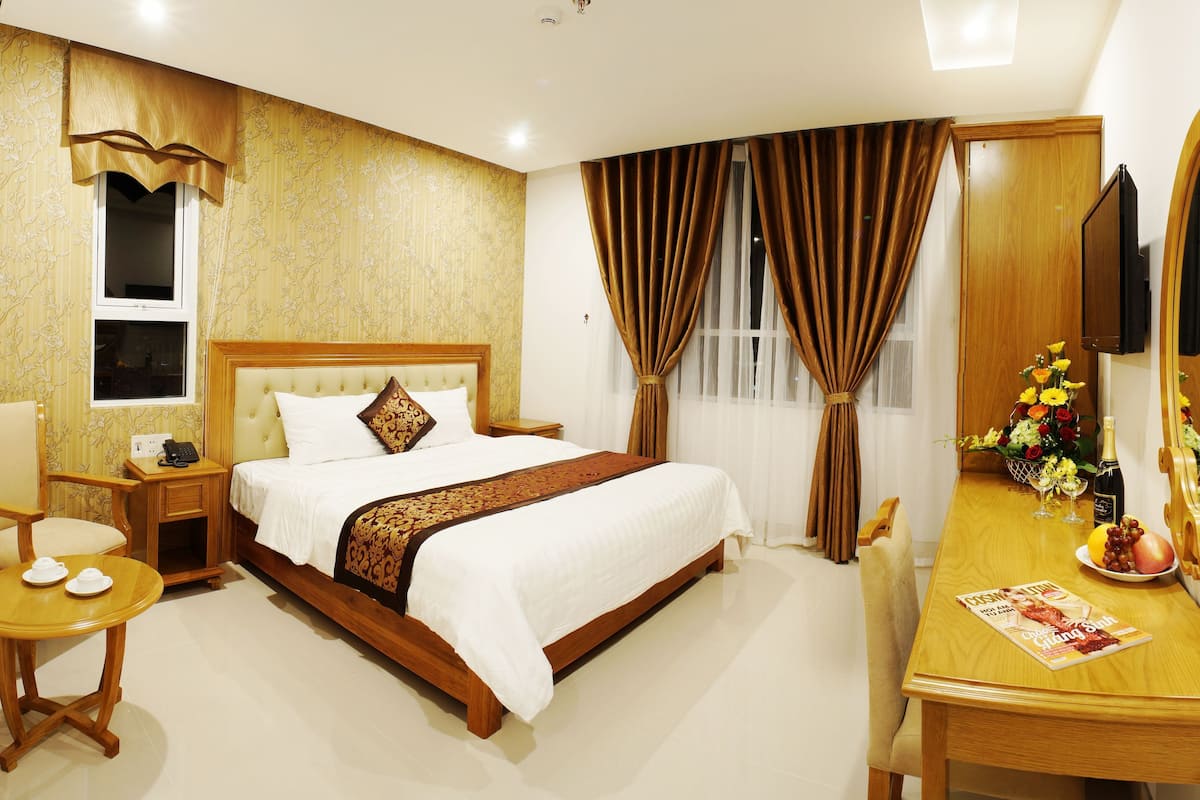 A hotel room with a bed, bedside table, chair, and a desk with a mirror and flowers.