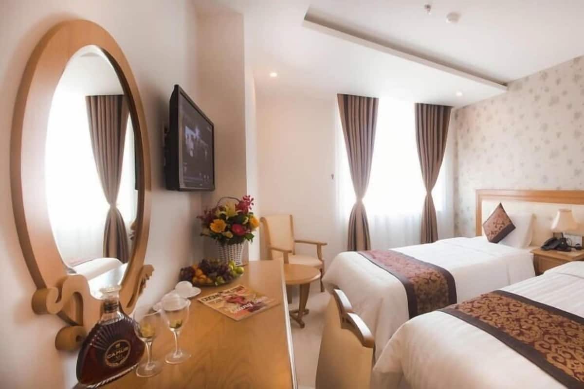 A hotel room with two beds, a desk with a bottle of liquor, glasses, and a magazine, a mirror, and a TV on the wall.