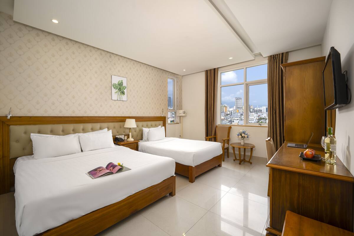 A hotel room with two beds, a desk, a chair, a small table, a TV, and a window with a city view.