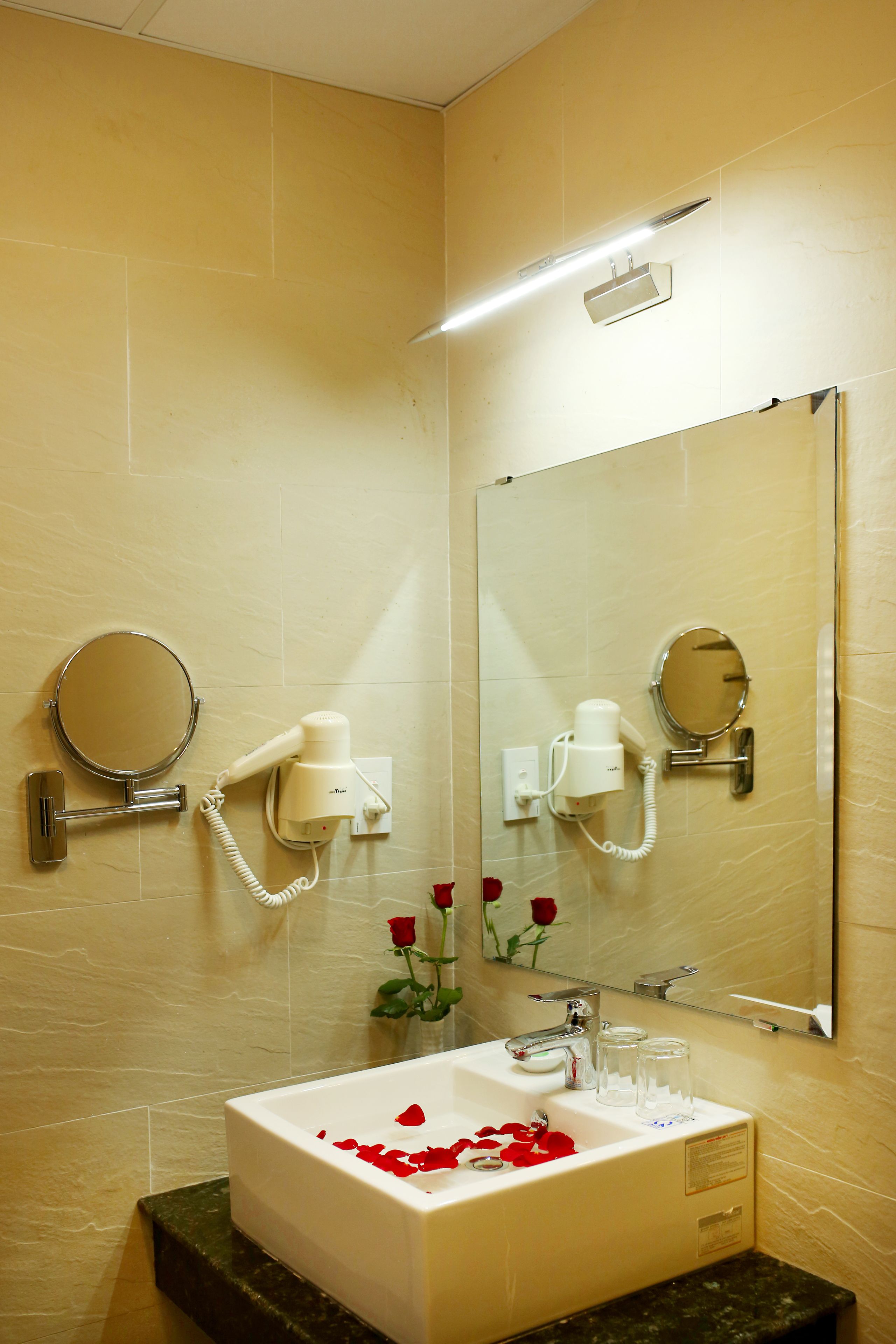 A bathroom with a sink, mirror, hairdryer, and a vase with roses.