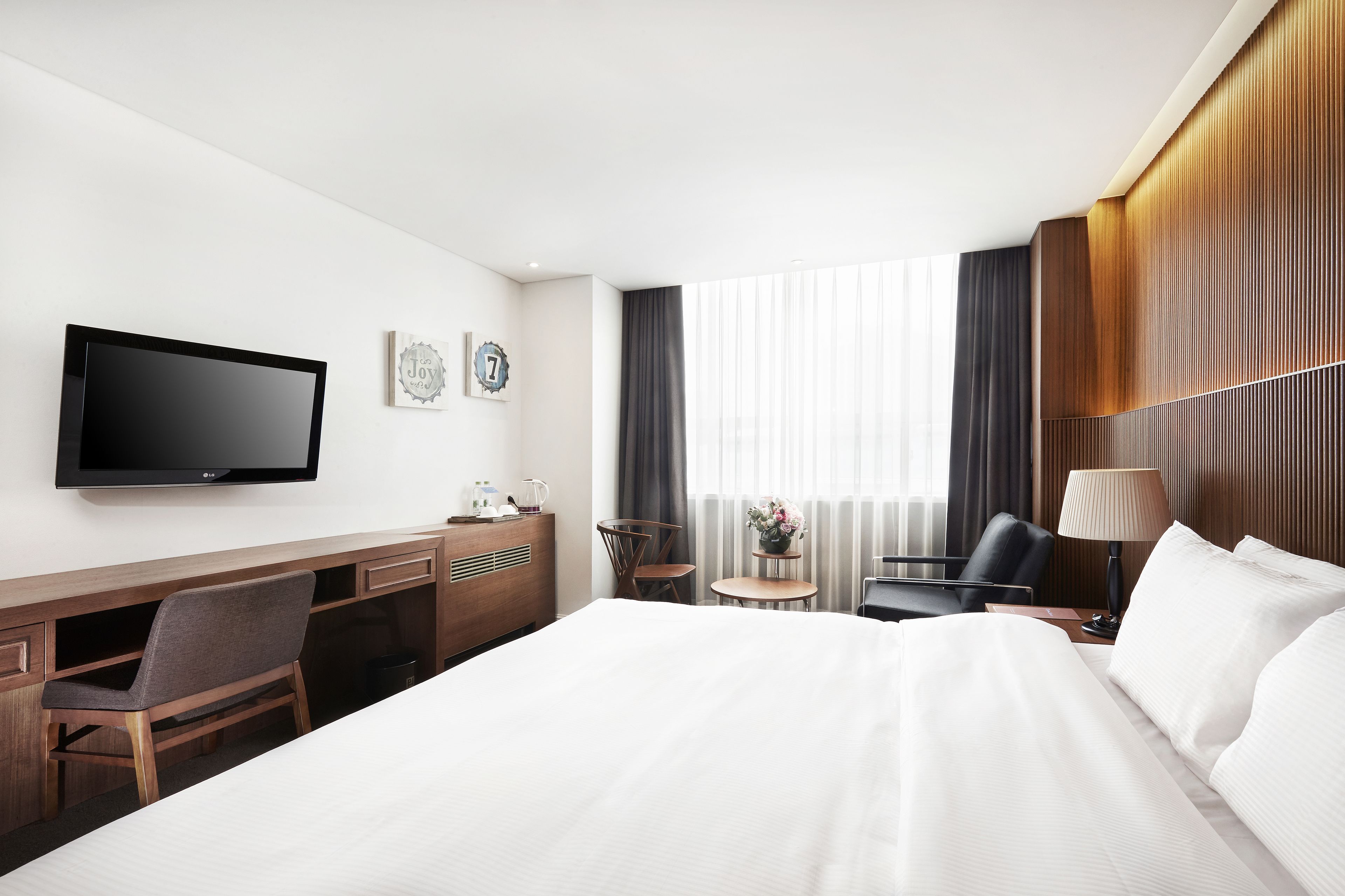 A hotel room with a bed, desk, chair, TV, and a small table with a lamp.