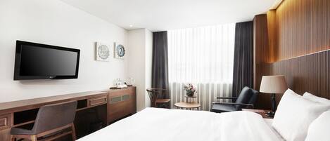 A hotel room with a bed, desk, chair, TV, and a small table with a lamp.