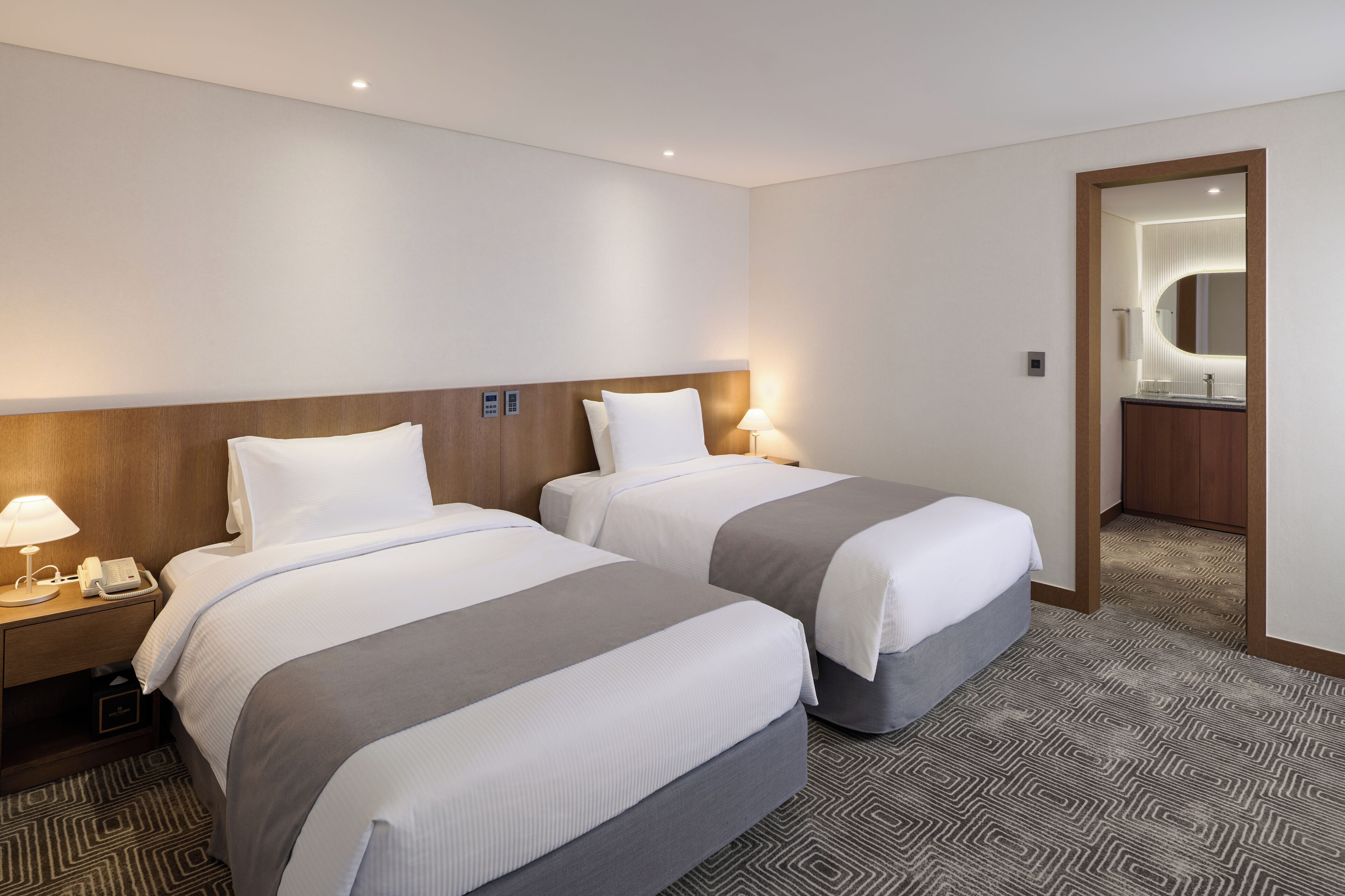 Habitación de hotel con dos camas, una mesita de noche con lámpara, un espejo y un baño visible a través de una puerta abierta.