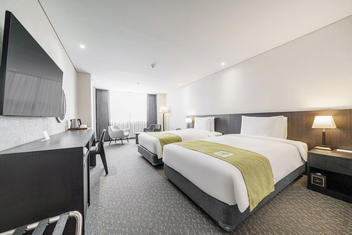 A hotel room with two beds, a desk, a chair, a TV, and a lamp.