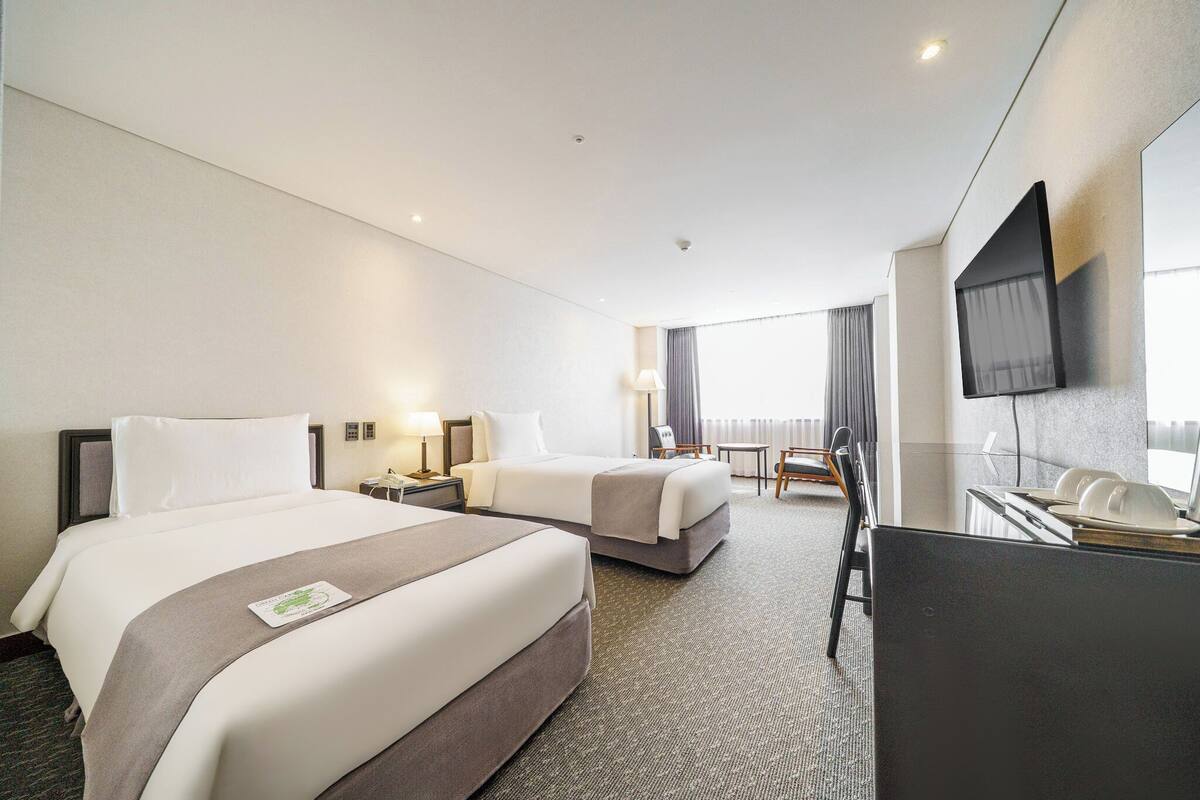 A hotel room with two beds, a desk, a chair, a TV, and a window with curtains.