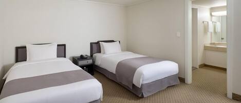 A hotel room with two beds, a nightstand with a phone, and a bathroom visible through an open doorway.