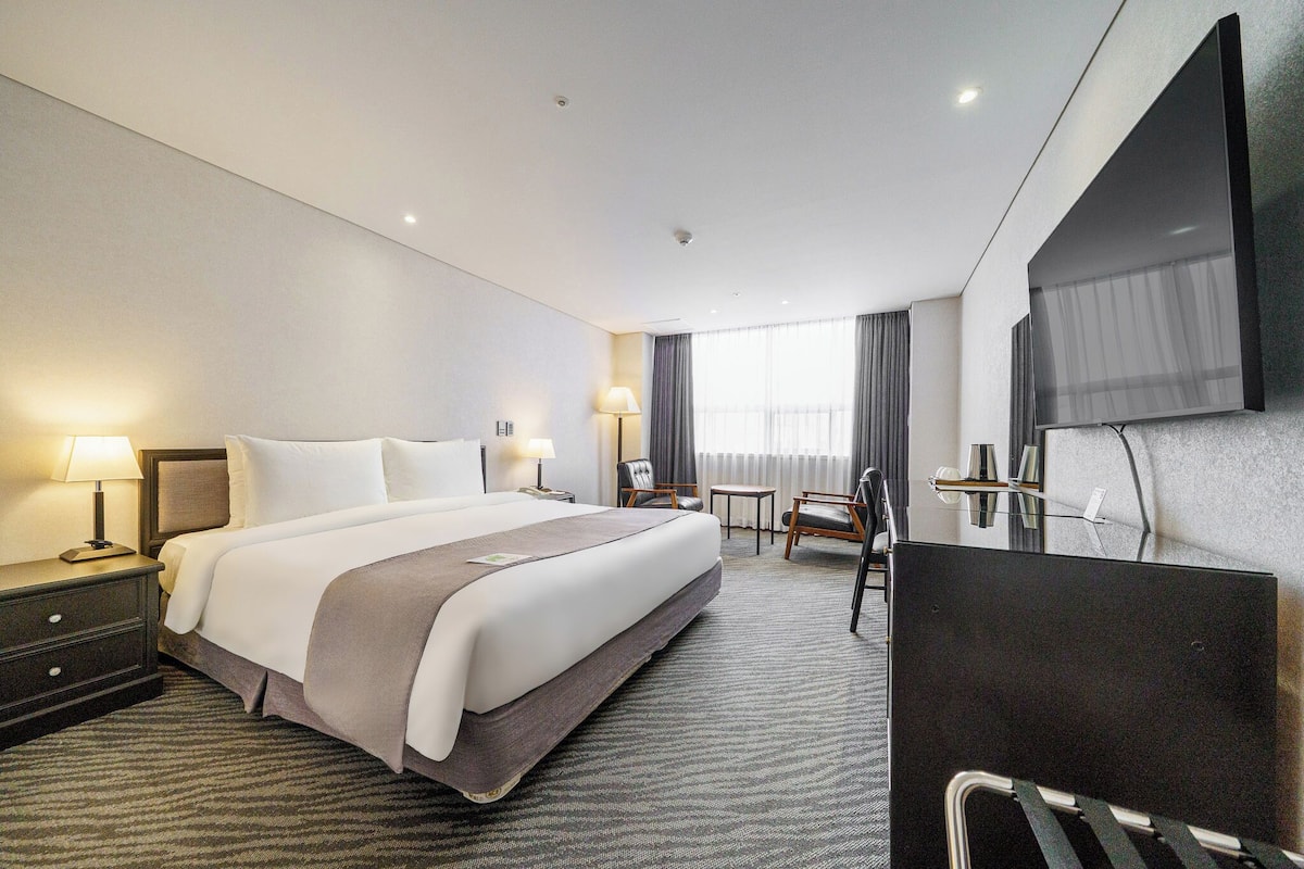 A hotel room with a large bed, a desk, a chair, a TV, and a window with curtains.