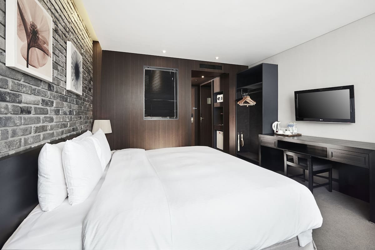 A modern hotel room with a large bed, a desk, a TV, and a brick wall.