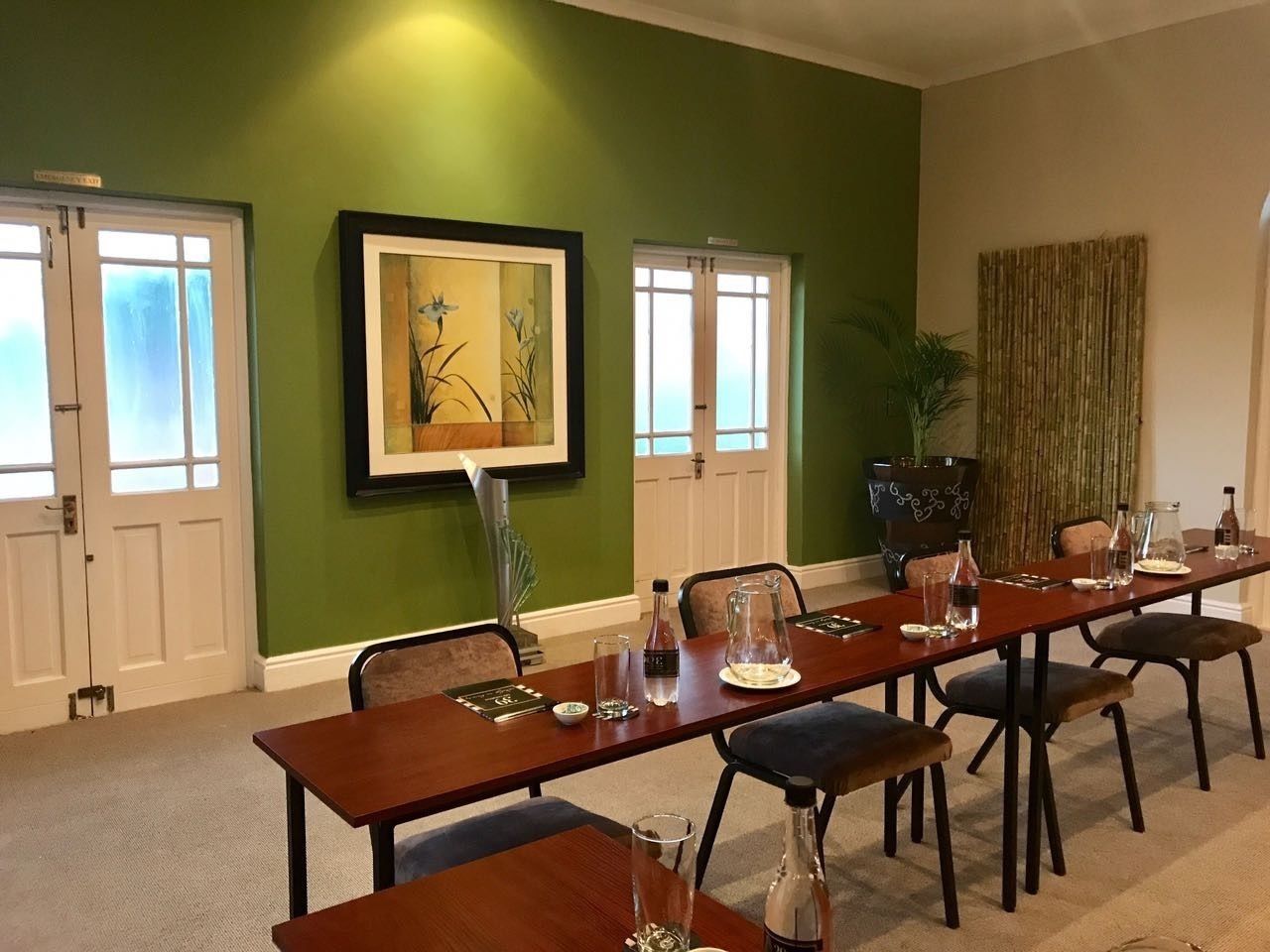Un comedor con una larga mesa de madera dispuesta para seis personas, flanqueada por dos puertas blancas, un cuadro enmarcado y una planta en maceta grande.