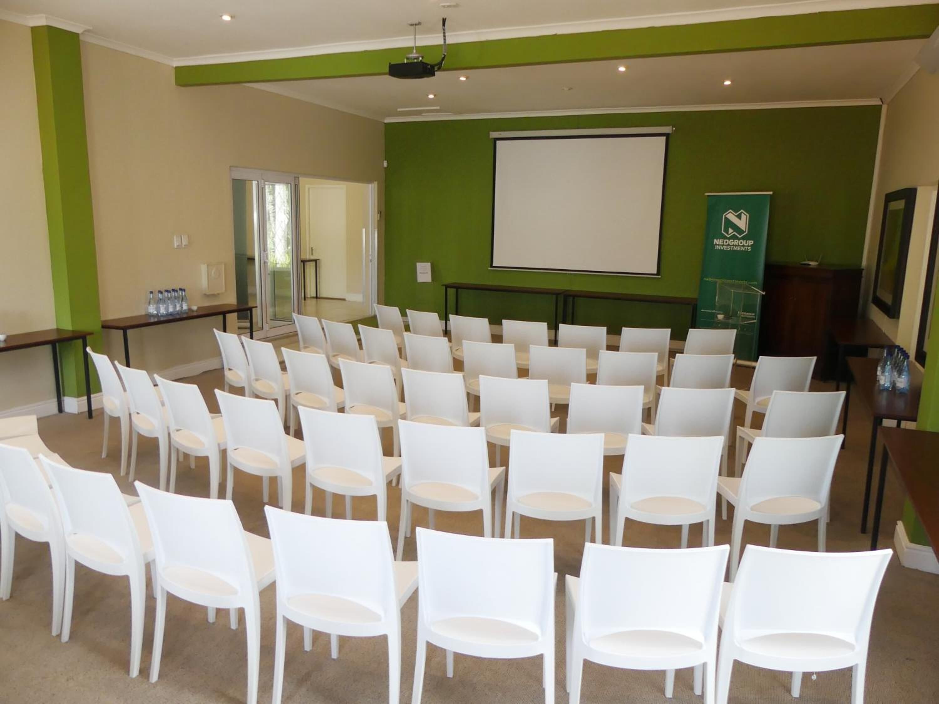A conference room with rows of white chairs, a projector screen, and a green wall with a logo.
