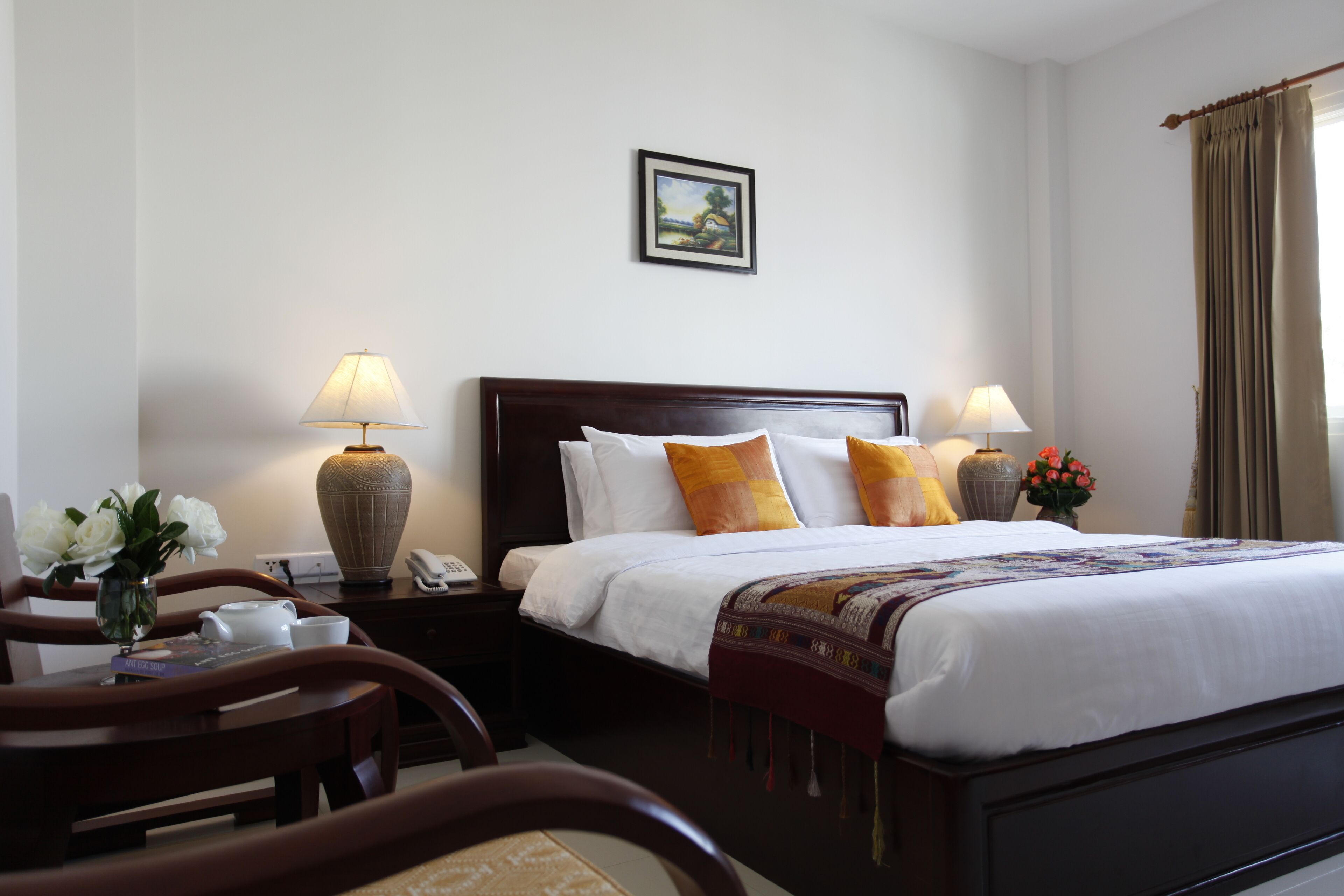 A hotel room with a large bed, two bedside tables, a desk with a chair, a vase of flowers, and a painting on the wall.