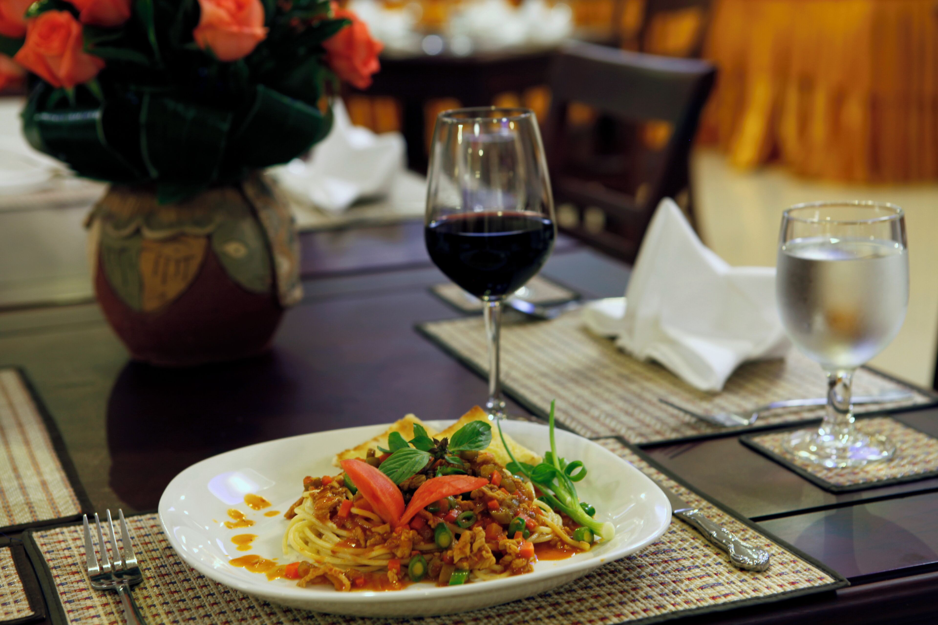Una mesa de comedor con un plato de espagueti, una copa de vino tinto y un florero con flores.