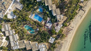 Vue aérienne d’un complexe hôtelier comprenant plusieurs bâtiments, une piscine centrale et une zone de plage.