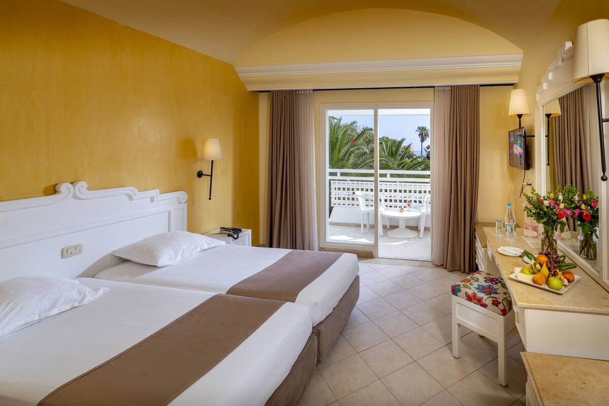Ein Hotelzimmer mit zwei Betten, einem Schreibtisch mit Obstschale, einem Spiegel und einem Balkon mit weißem Zaun und Palmen.