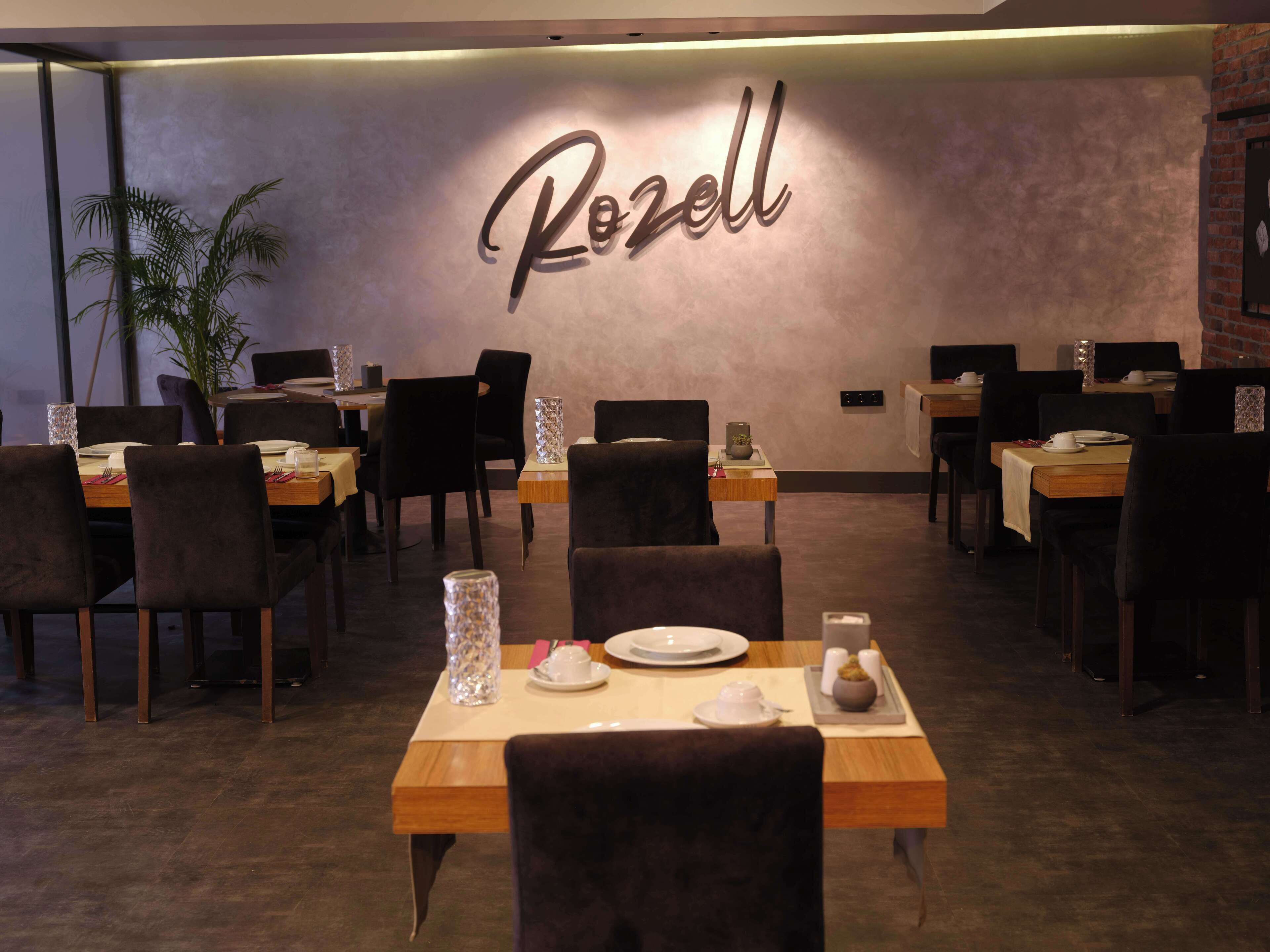 A restaurant interior with tables set for dining, a wall-mounted sign reading "Rozell," and a potted plant in the corner.