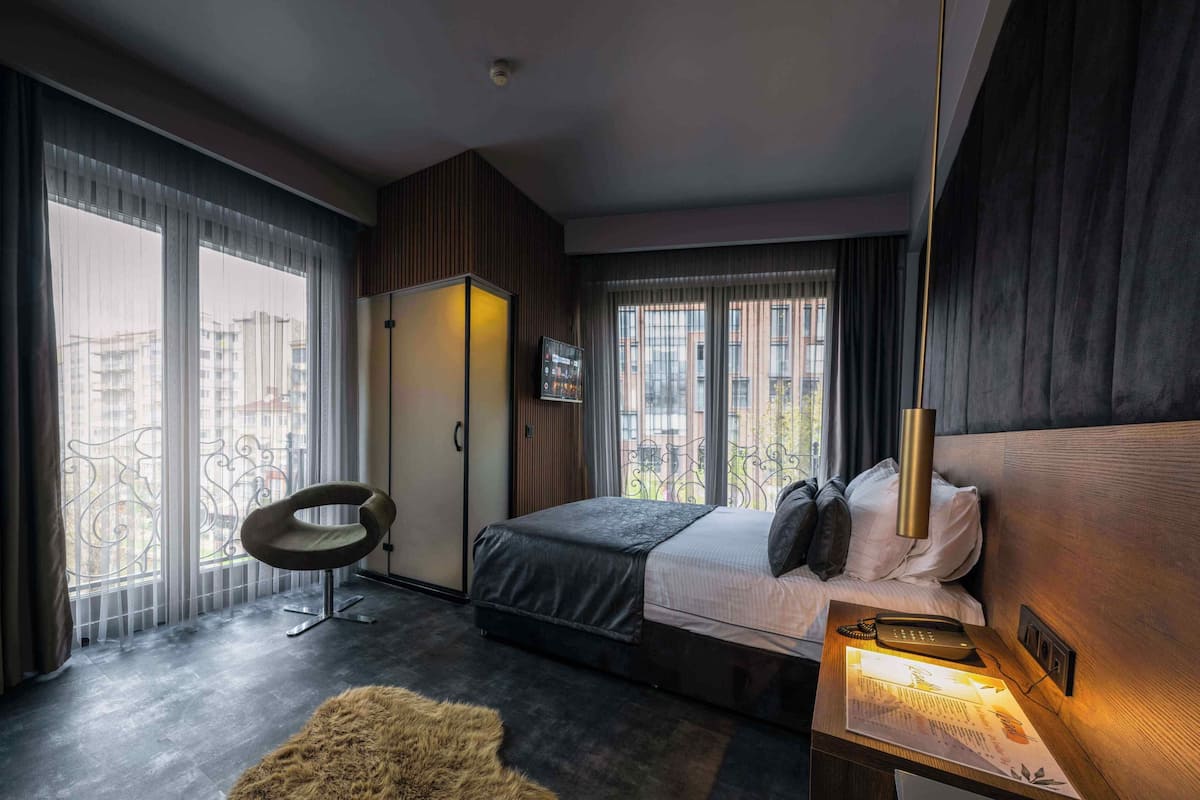 Een moderne hotelkamer met een bed, nachtkastje, stoel, kledingkast en grote ramen met uitzicht op gebouwen.