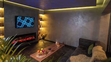 A modern living room with a flat-screen TV, a fireplace, a wooden coffee table with fruit and a glass of wine, and a cozy sofa with a fur throw.