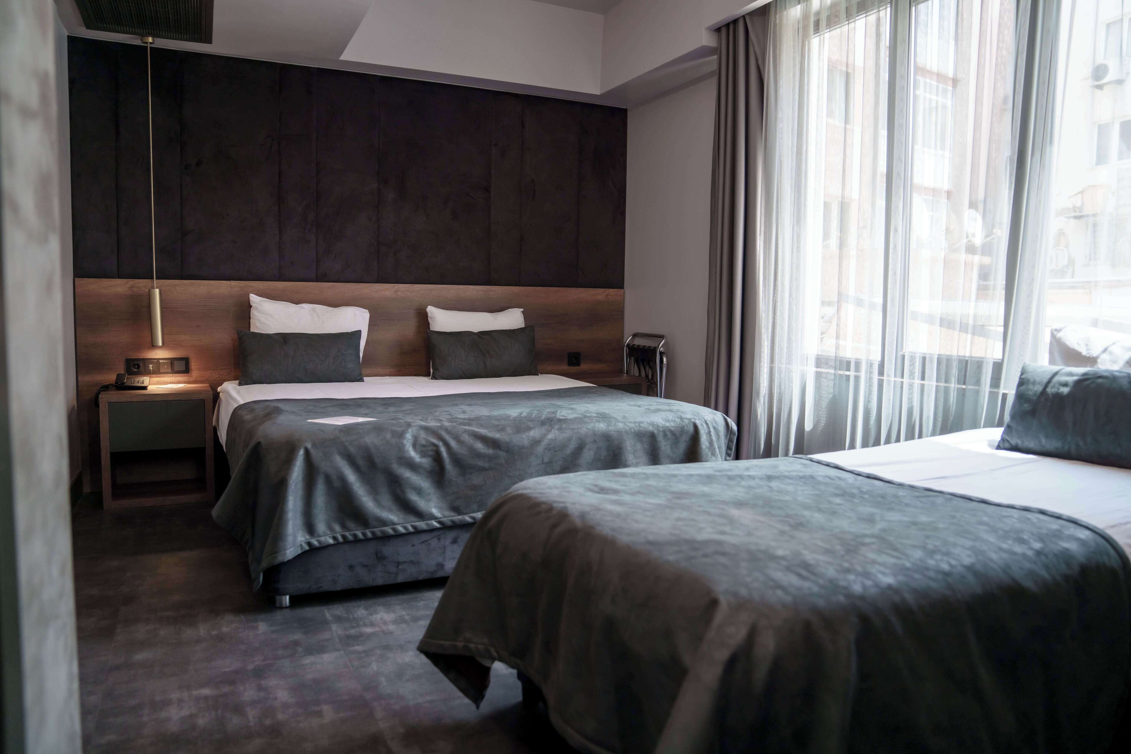 Una habitación de hotel moderna con dos camas, cabecero de madera oscura, mesitas de noche con lámparas y un amplio ventanal con cortinas.