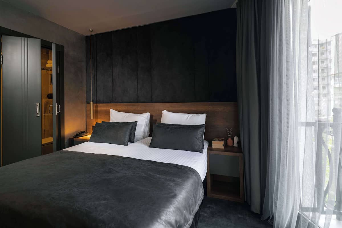 Een moderne hotelkamer met een groot bed, een houten hoofdbord, een nachtkastje met een lamp en uitzicht op de stad door dunne gordijnen.