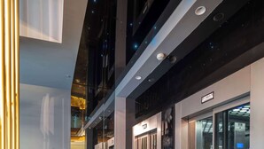 Un couloir d’hôtel moderne avec des portes en verre, un écran numérique indiquant l’étage de l’ascenseur et des plantes en pot.