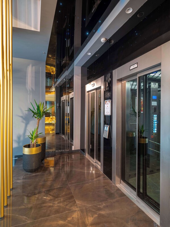 A modern hotel corridor with glass doors, a digital elevator display, and potted plants.