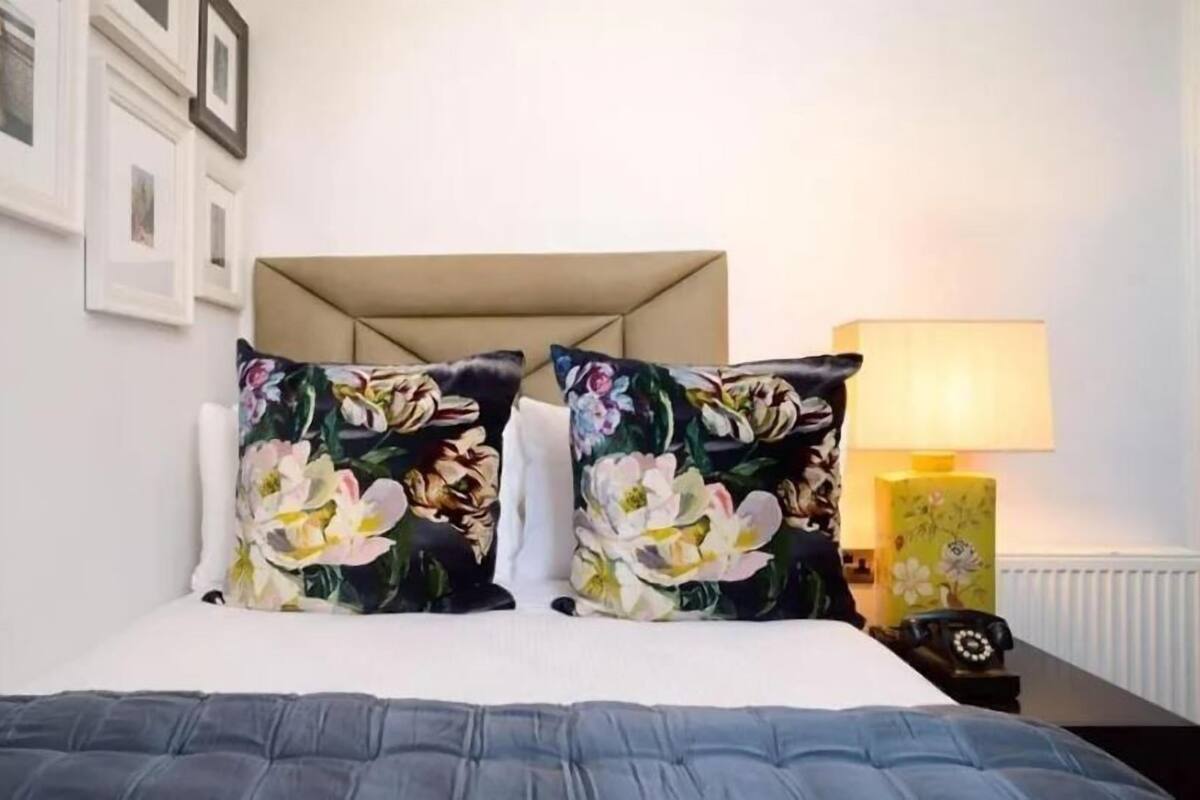 A bedroom with a bed, two floral pillows, a bedside lamp, and a framed picture on the wall.