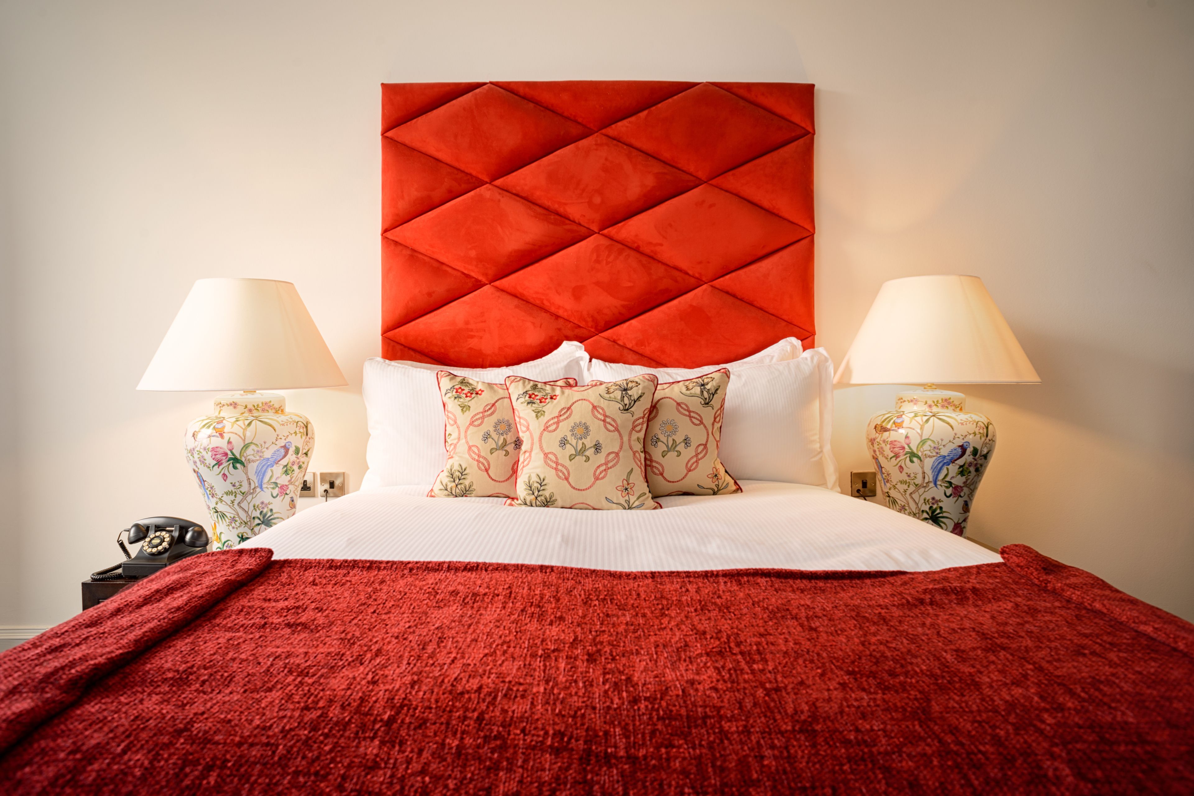 Une chambre d’hôtel avec une tête de lit rouge, des lampes à motifs floraux et un plaid rouge.