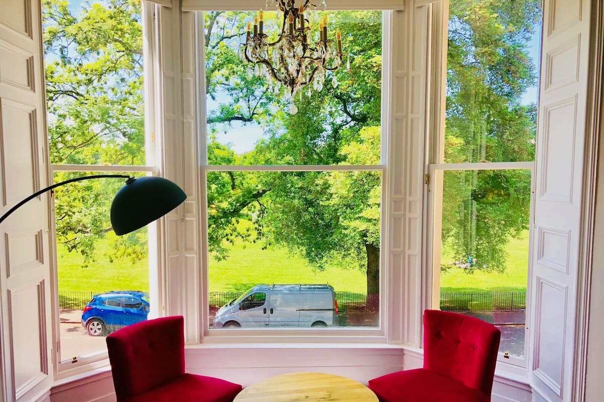A room with two red chairs, a round wooden table, and a large window with a view of a green park and a white van.