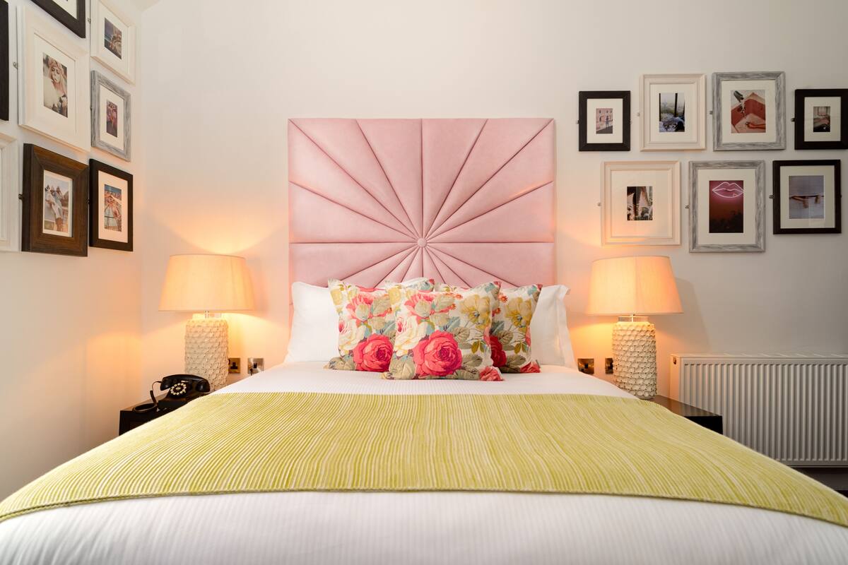 A neatly arranged bedroom with a large bed, two bedside lamps, a decorative headboard, and a series of framed pictures on the wall.
