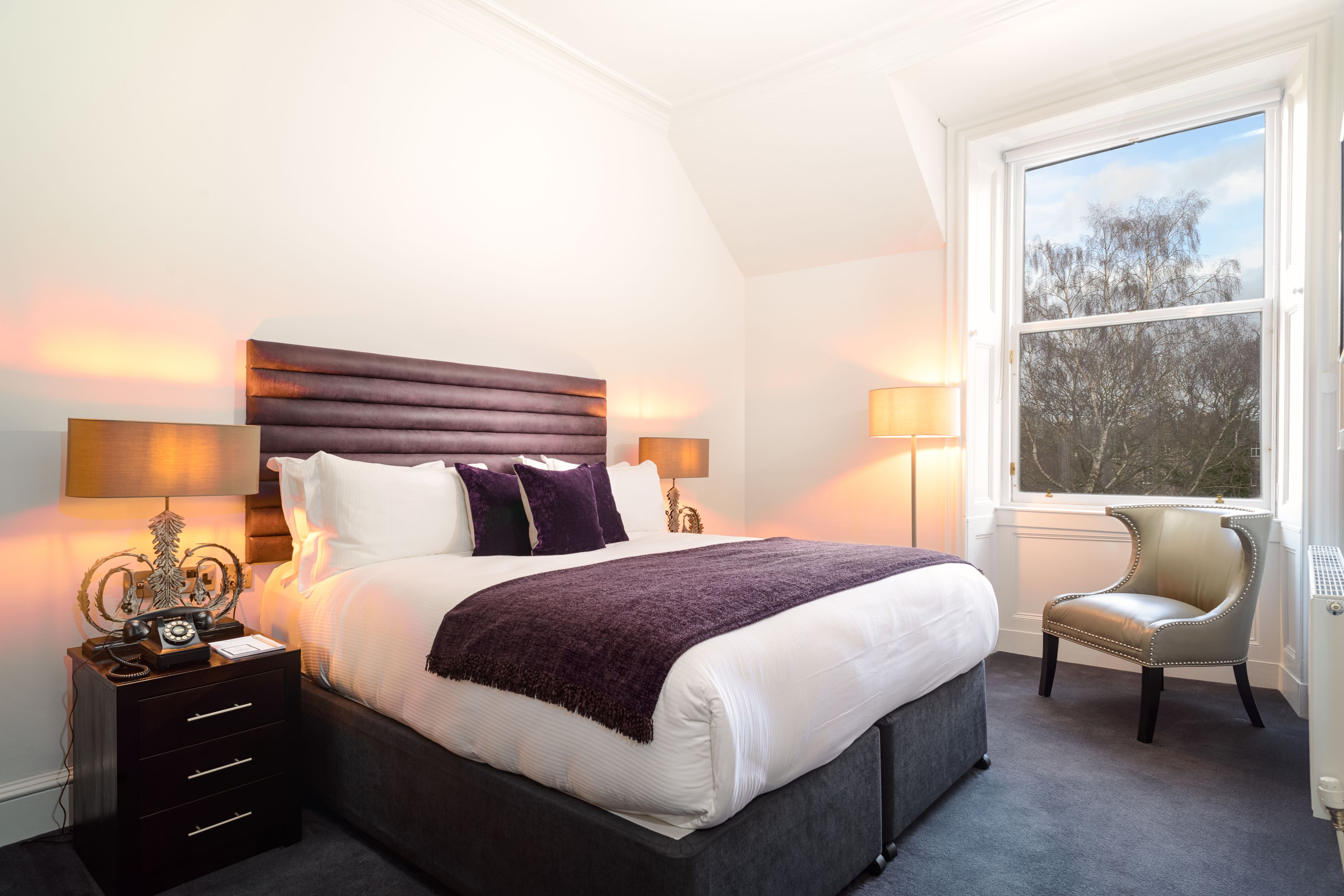 A neatly made bed with a headboard, bedside lamps, a chair, and a window with a view of trees.