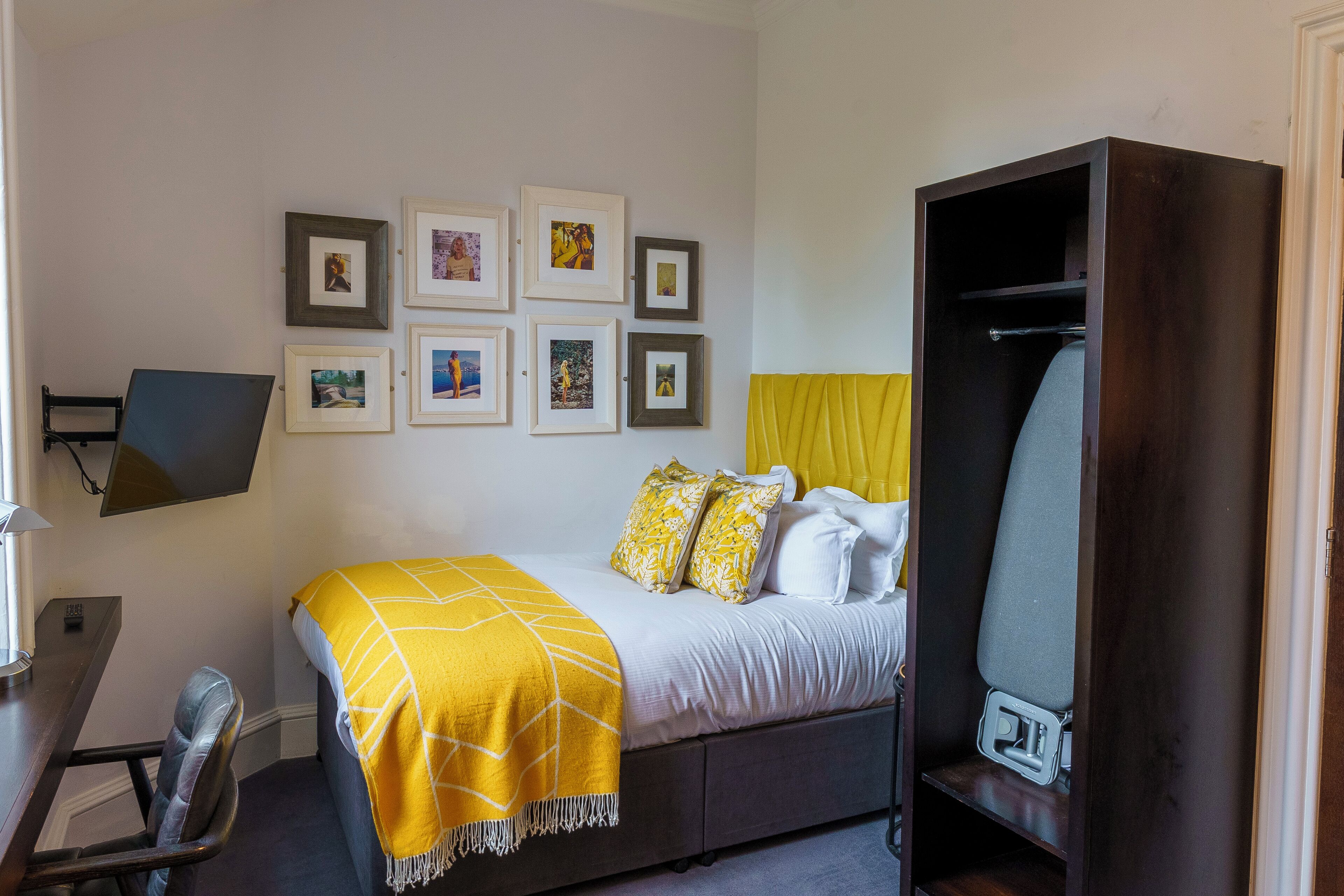 A bedroom with a bed, a desk, a chair, a wardrobe, and framed pictures on the wall.