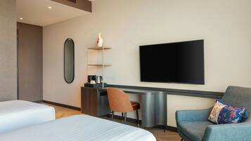 Una habitación de hotel moderna con dos camas, un escritorio con televisor de pantalla plana, una silla y una pequeña estantería con un jarrón.