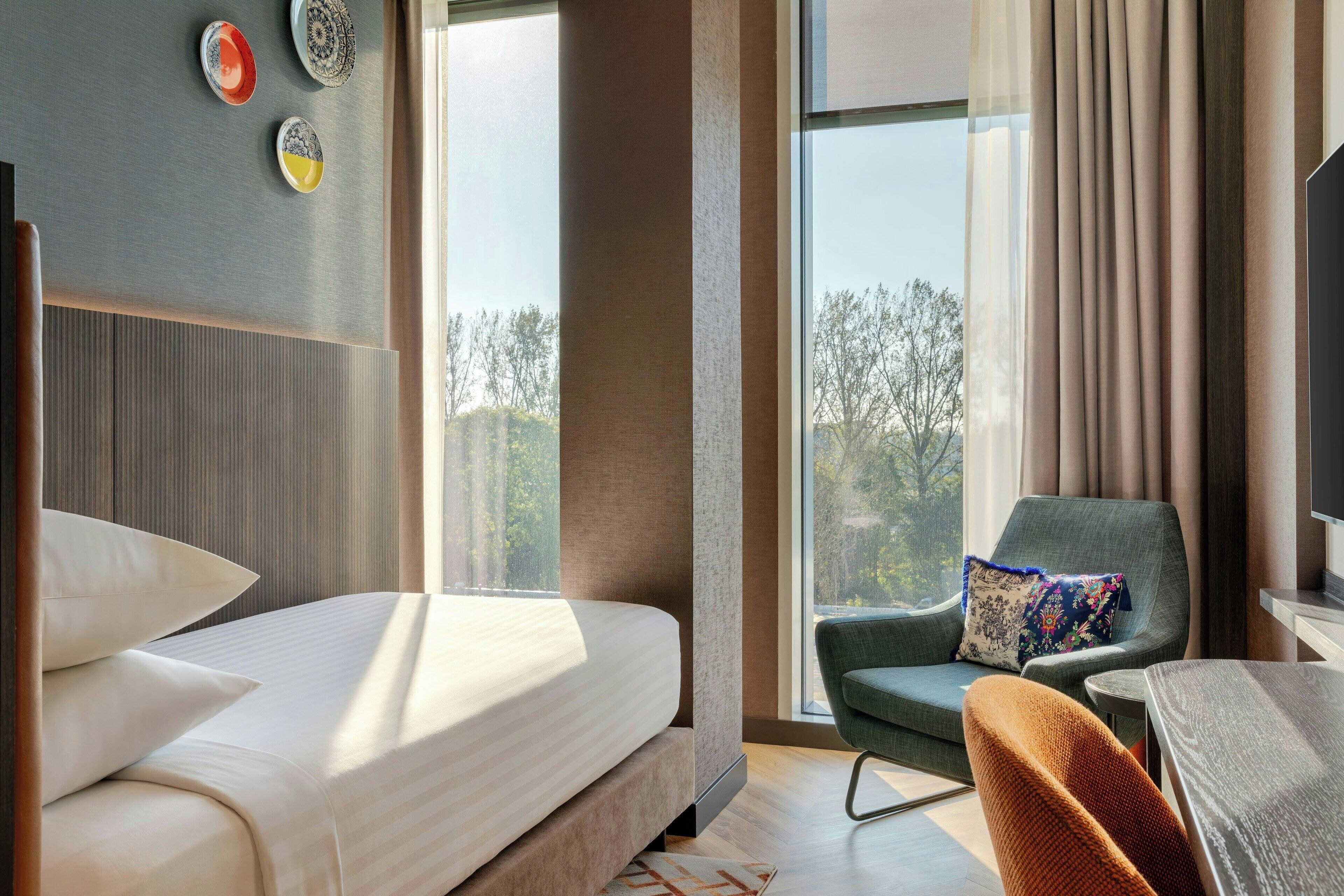Habitación de hotel moderna con una cama grande, un sillón cómodo, una mesita y vistas a árboles por la ventana.