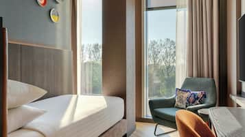 A modern hotel room with a large bed, a comfortable armchair, a small table, and a view of trees through the window.
