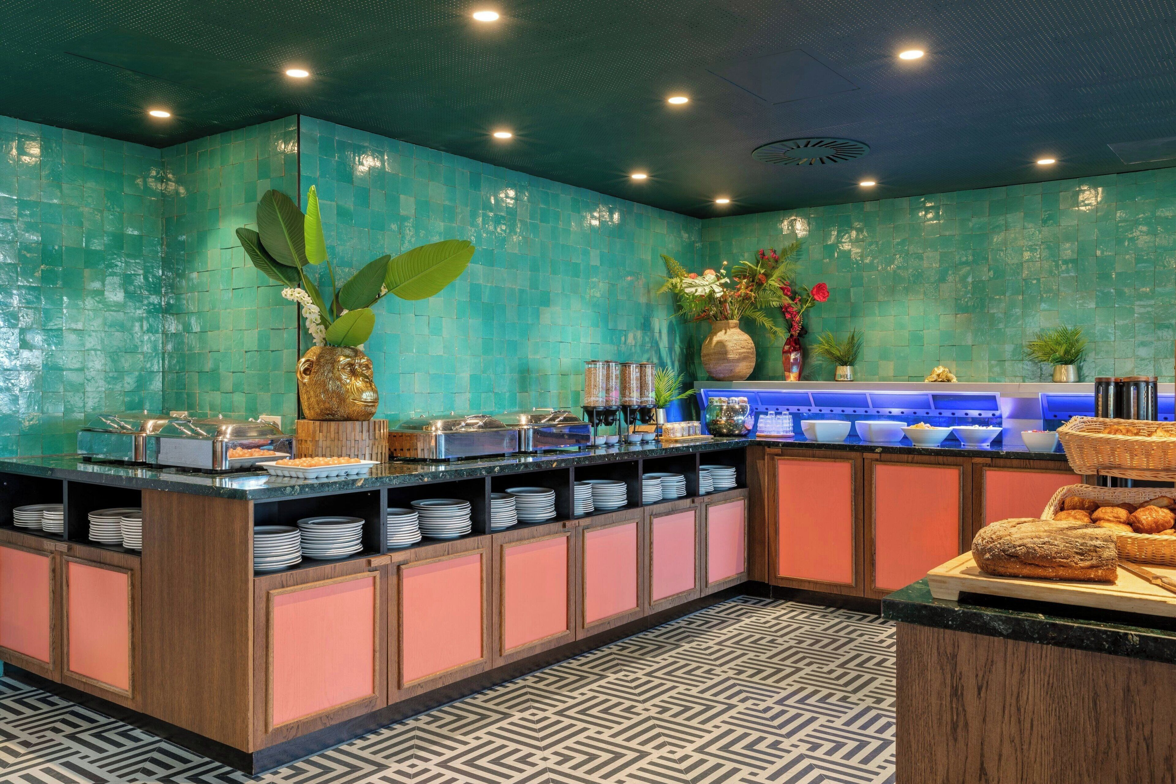 Un área de bufé con una pared de azulejos color turquesa, piso estampado y diversas opciones de comida exhibidas en estantes.
