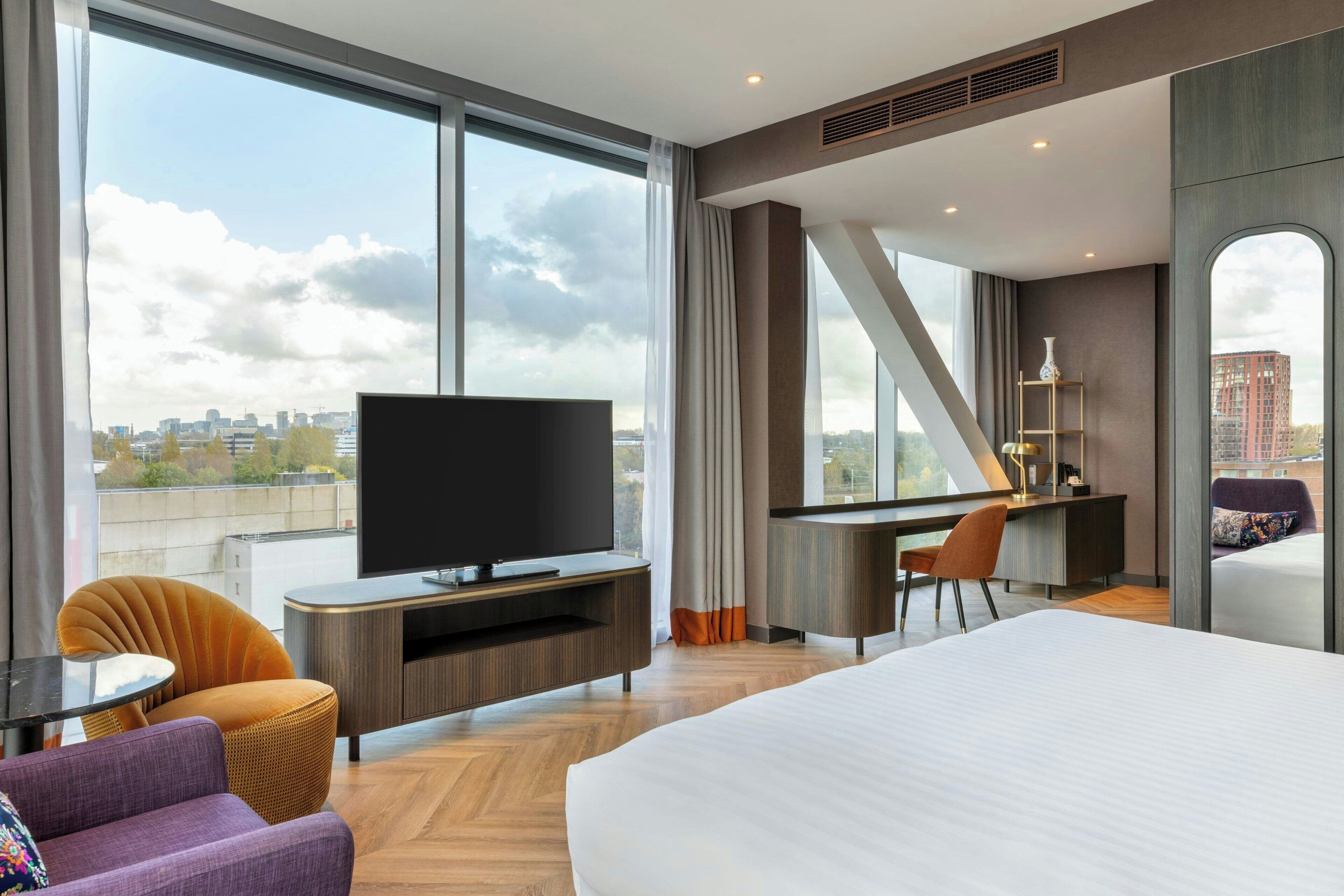 Een moderne hotelkamer met een groot bed, een flatscreen televisie, een bureau met stoel, en uitzicht op de stad door grote ramen.