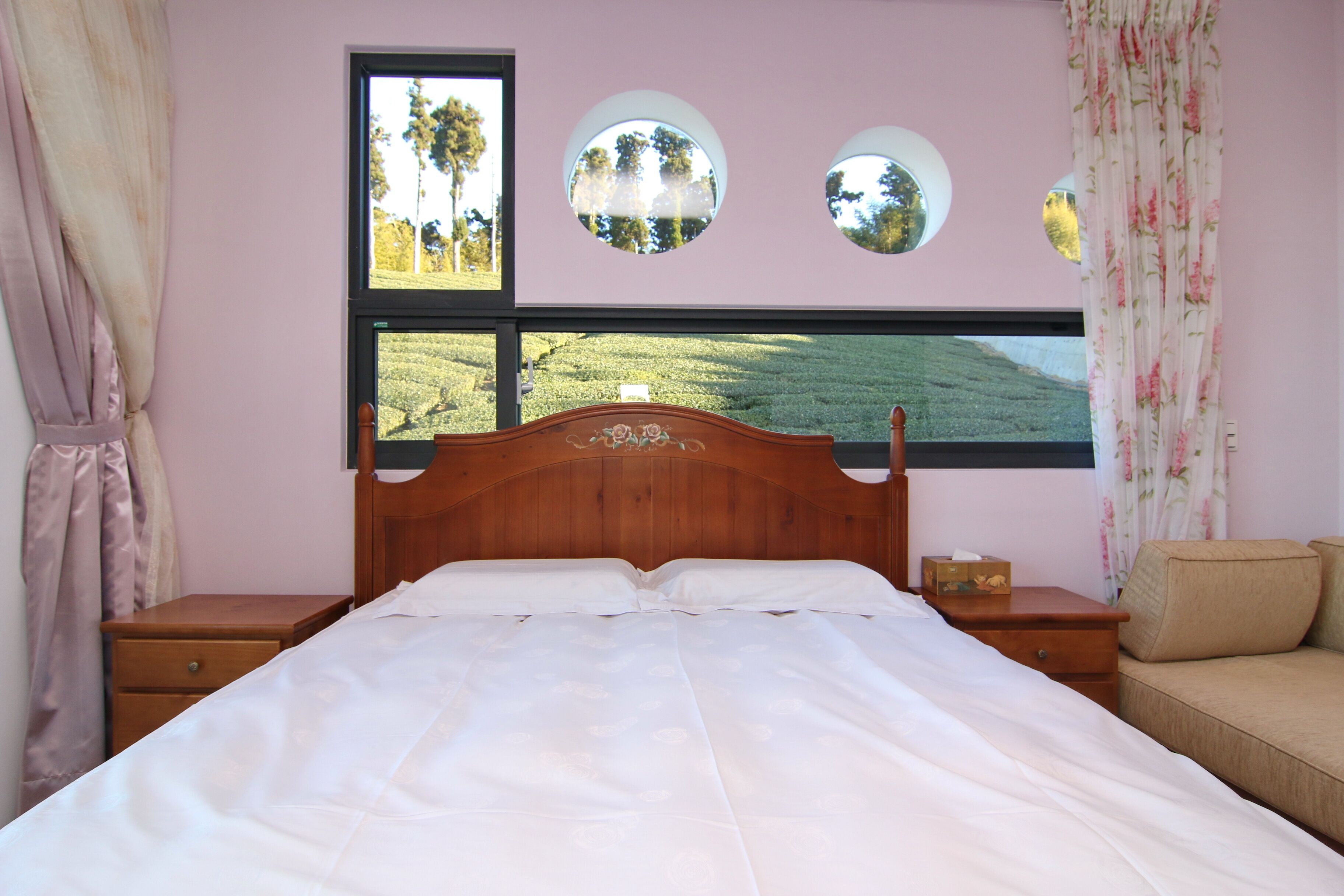 A bedroom with a wooden headboard, a large window with a view of a green landscape, a bedside table with a lamp, and a sofa.