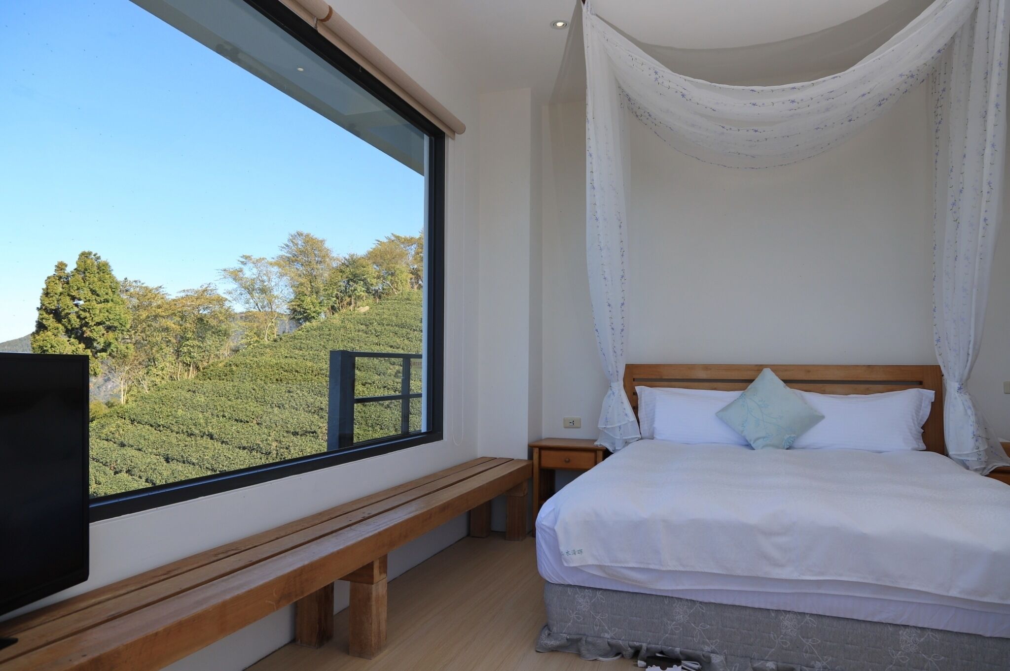 A bedroom with a large window, a wooden bench, a bed with white bedding, and a nightstand.