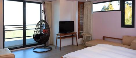 A modern hotel room with a large bed, a television on a wooden stand, a hanging chair, and large windows offering a view of the outdoors.