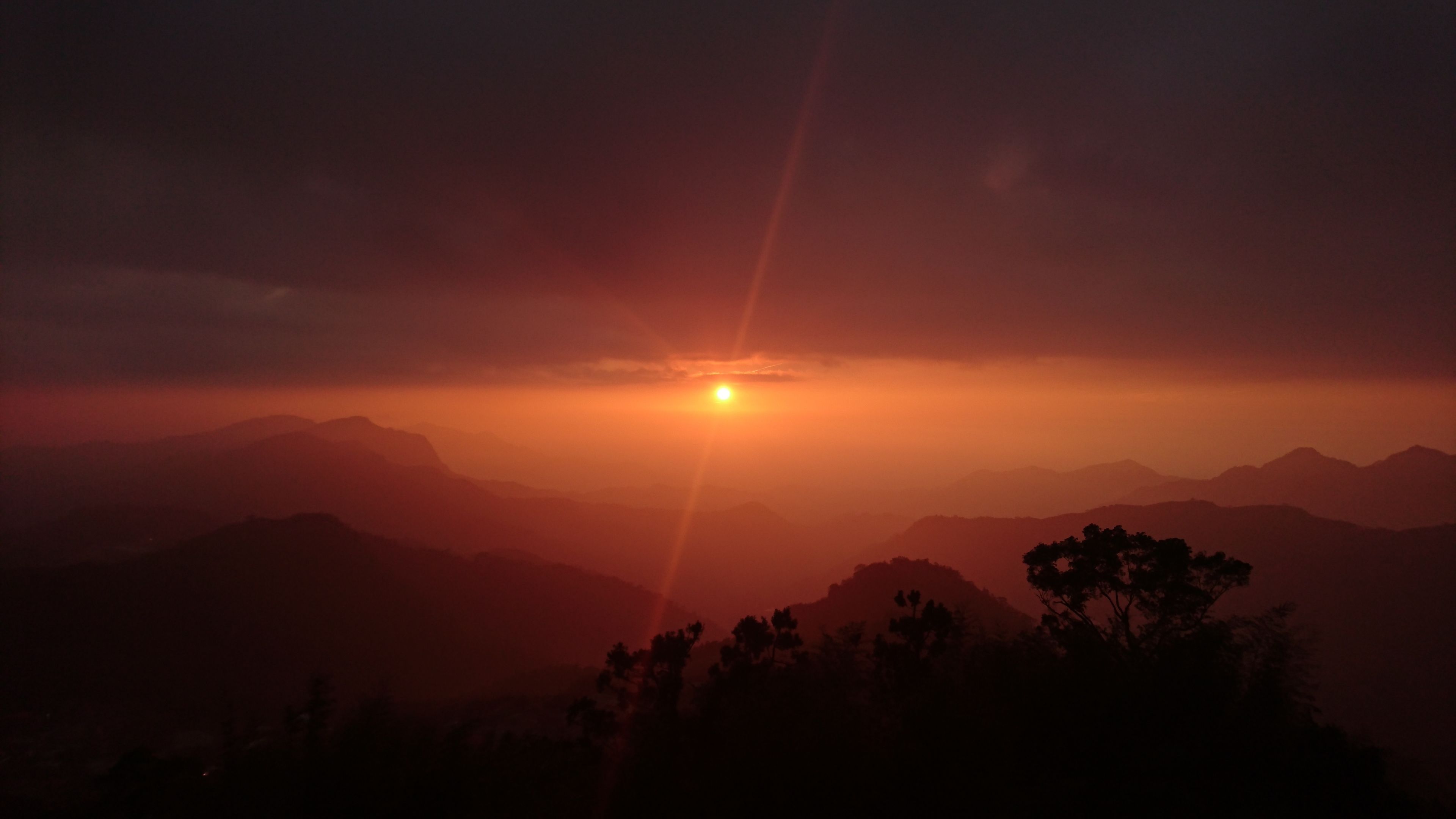 Ein Sonnenuntergang über einer Bergkette, im Vordergrund Silhouette von Bäumen.