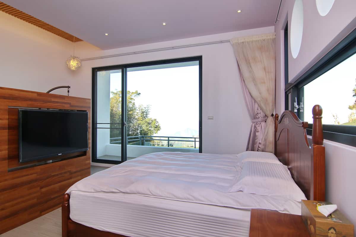 Une chambre à coucher avec un lit, une télévision, une commode en bois et une grande fenêtre donnant sur les arbres.