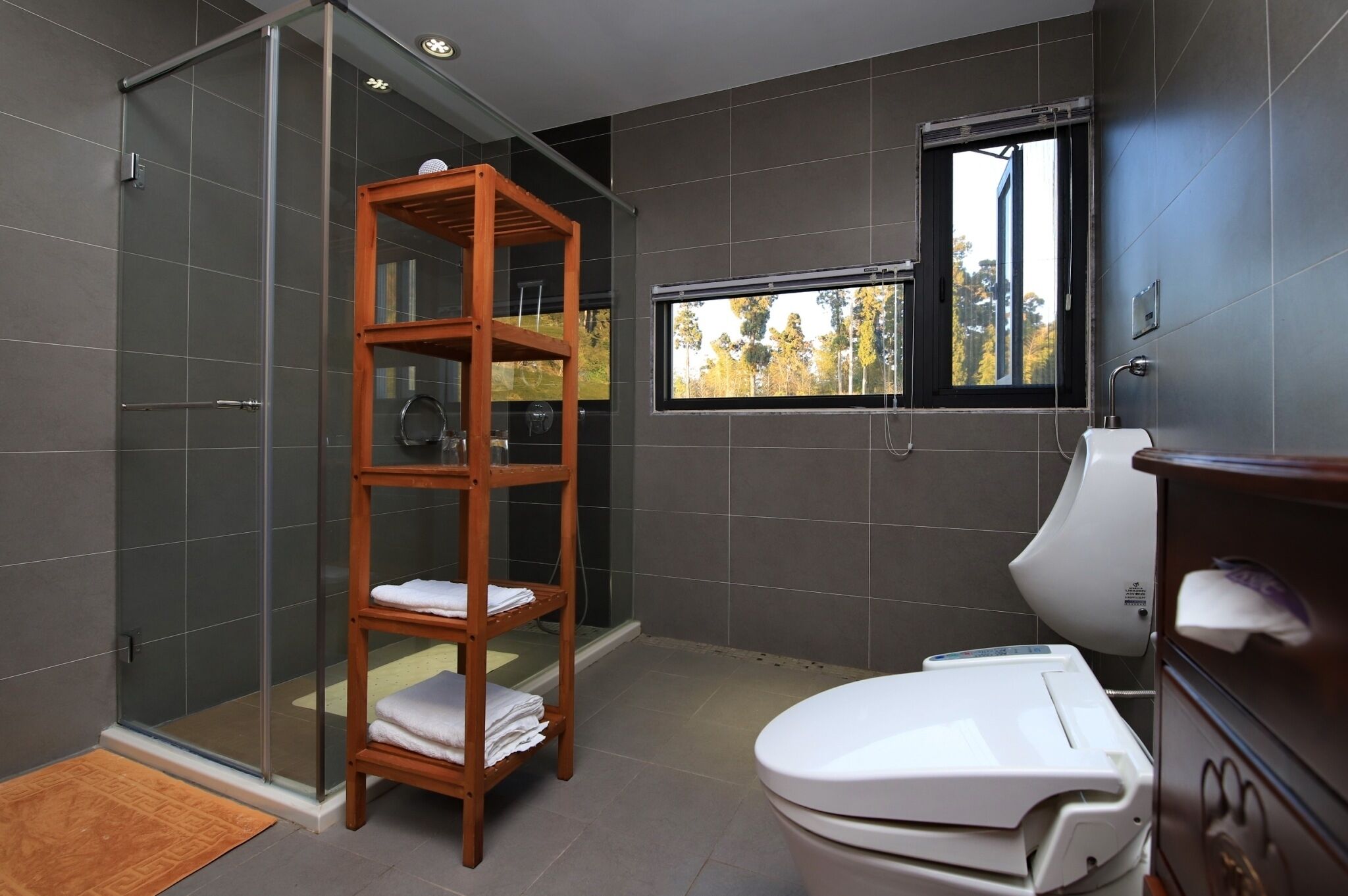 Un baño moderno con cabina de ducha de vidrio, repisa de madera con toallas, inodoro y ventana con vistas a árboles.