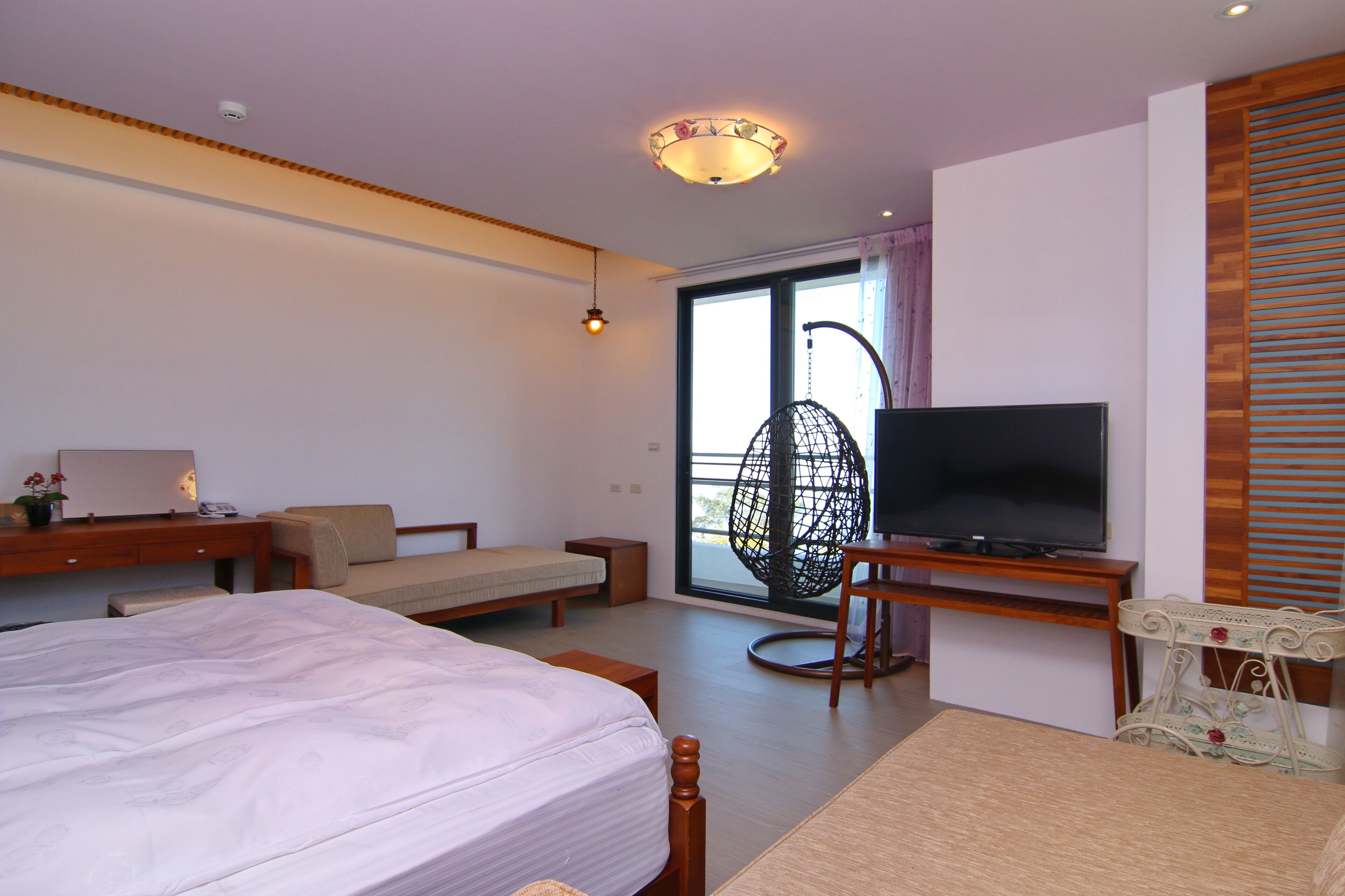 A modern hotel room with a large bed, a television, a desk, and a balcony with a wicker chair.