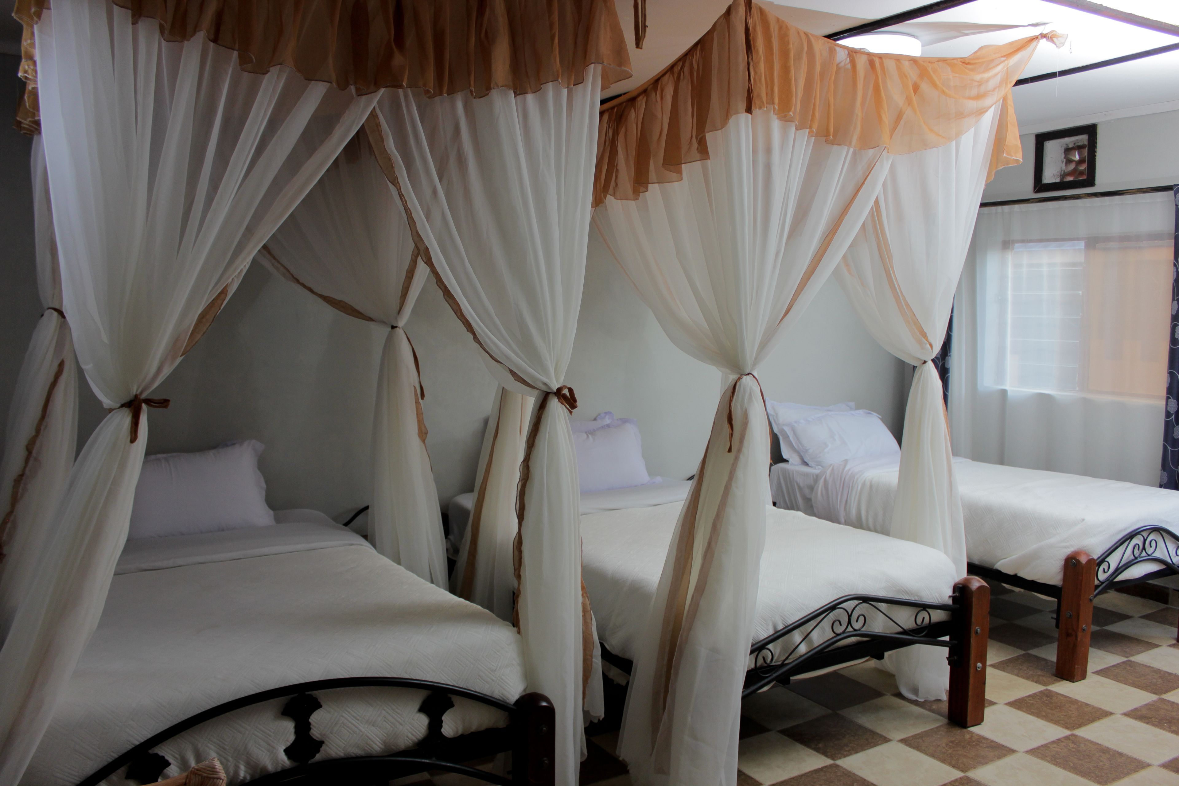 A room with four beds, each with a canopy bedspread and white bedding. The room has a checkered floor and a window with curtains.