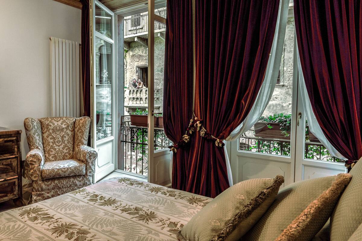 Una habitación con una cama, una silla, un ventanal con cortinas y vistas a un balcón.