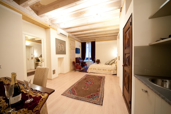 A well-lit room with a dining table set for two, a bed with a floral bedspread, a small kitchen area, and a hallway leading to another room. - La Corte di Giulietta Exclusive Suites (Verona)