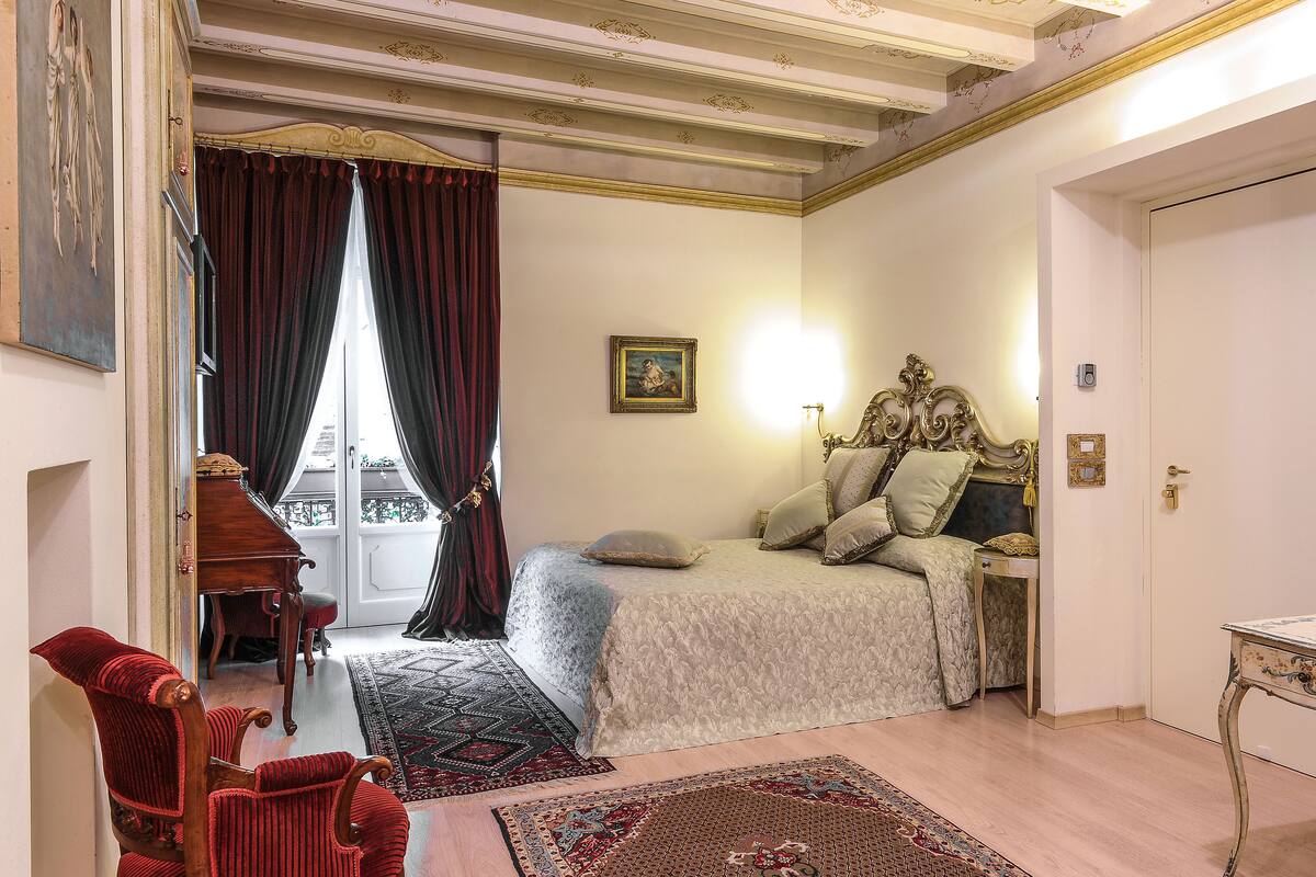 Una habitación con una cama, un escritorio y una silla. Hay una ventana con cortinas, un cuadro en la pared y una puerta.