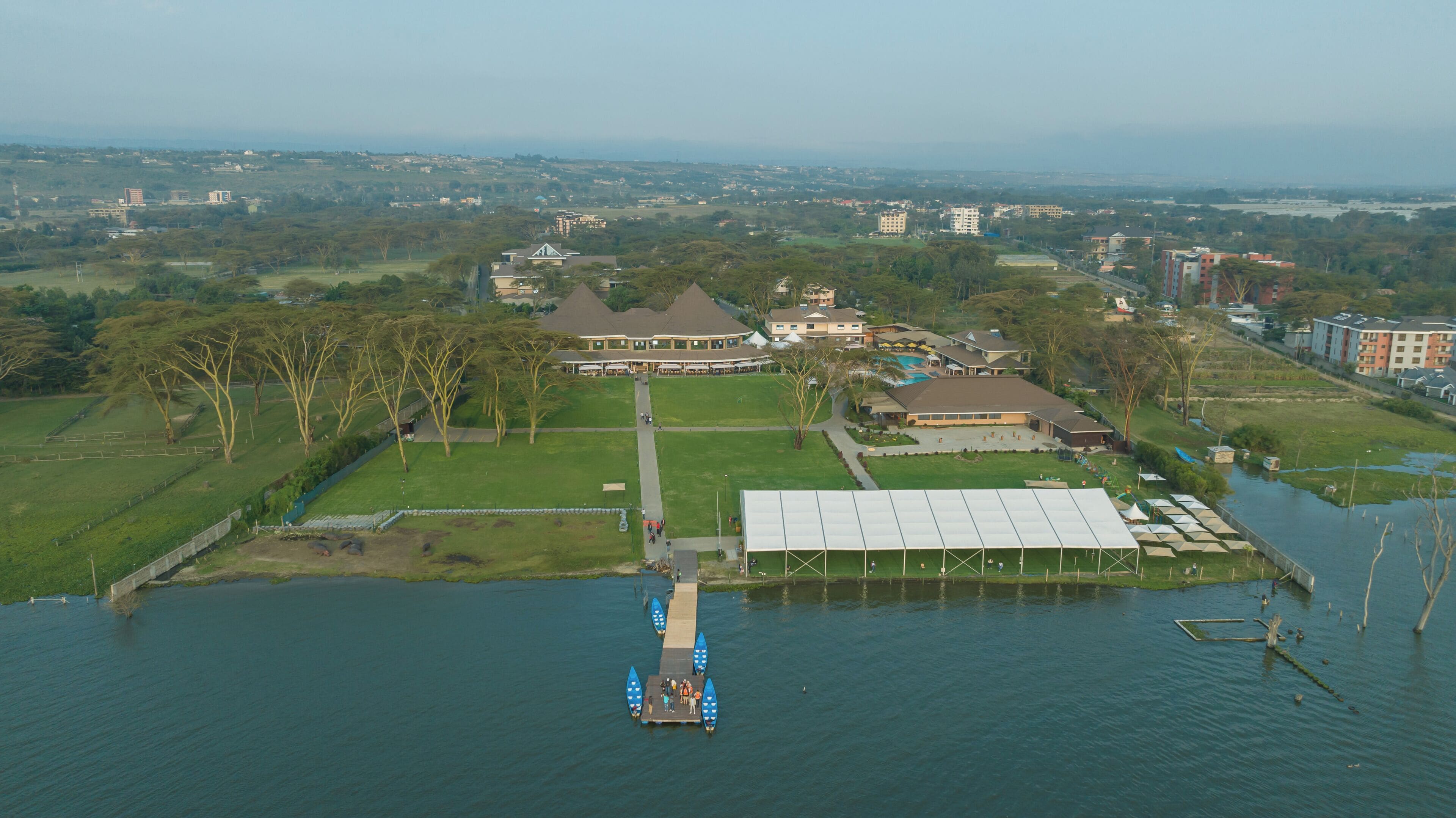 An aerial view of a lakeside property with a large building, a dock with boats, and surrounding greenery.