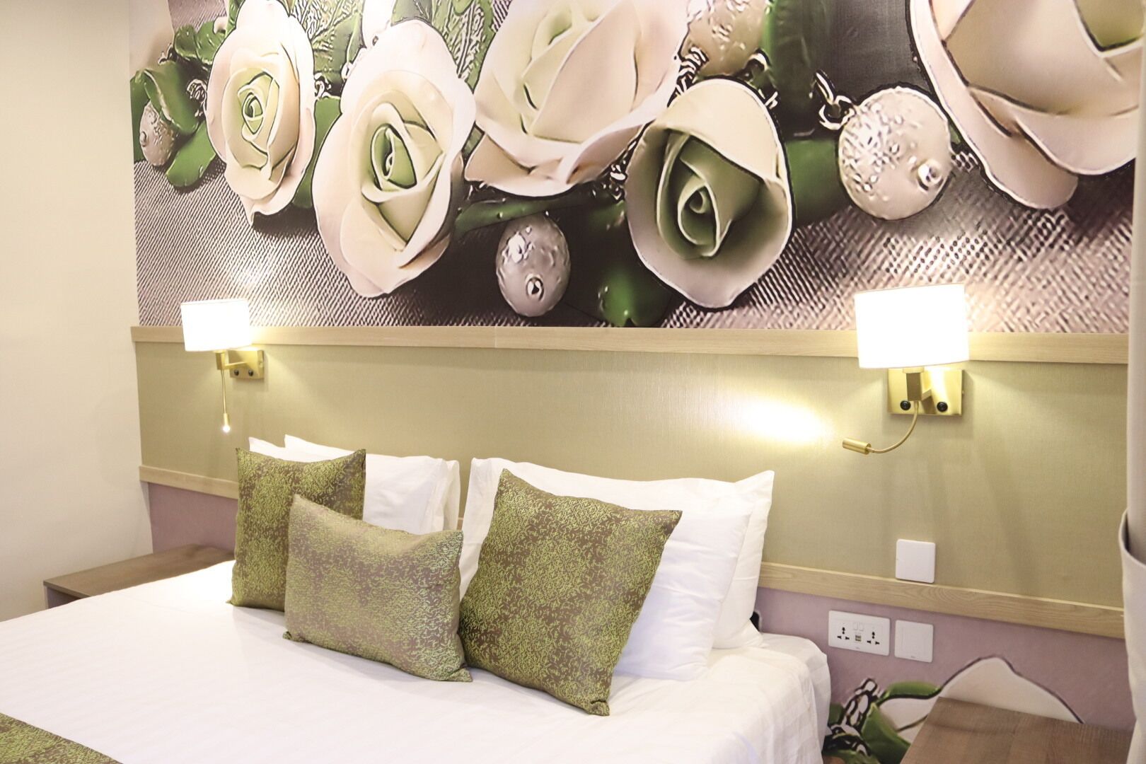 Une chambre d’hôtel avec un lit, deux oreillers, une lampe fixée au mur et une fresque murale à motifs floraux.