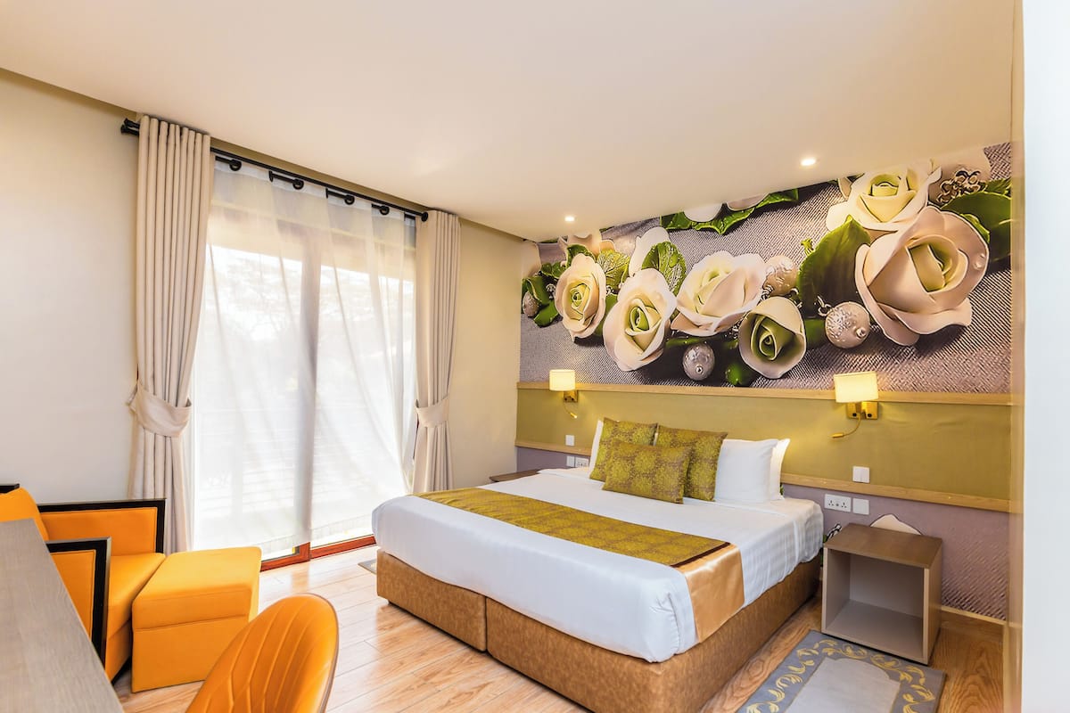 A hotel room with a bed, a seating area, a wall-mounted artwork of flowers, and a window with curtains.